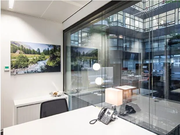 Een modern kantoor met een bureau, telefoon, lamp en een schilderij van een rivierlandschap, naast een glazen wand met uitzicht op een atrium.