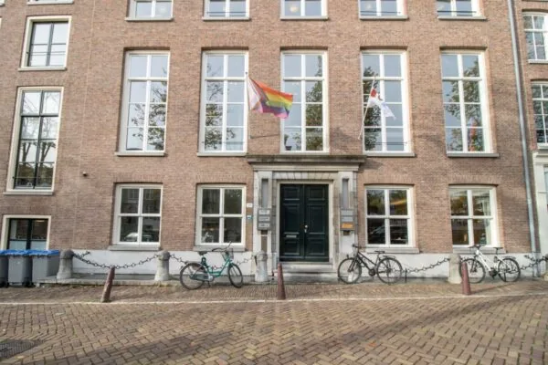Voorgevel van een kantoorpand aan de Nieuwe Herengracht 17 in Amsterdam met regenboog- en witte vlag, fietsen en grote ramen.