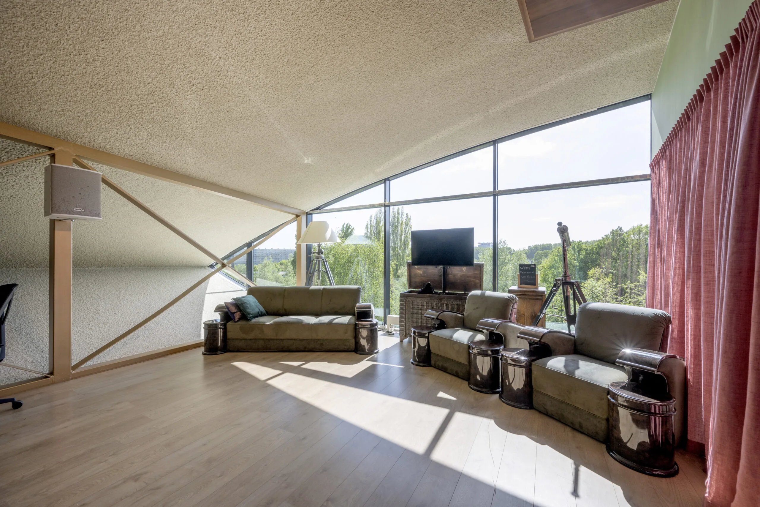 Een ruime woonkamer met grote ramen, moderne meubels en een telescoop met uitzicht op groene bomen buiten.