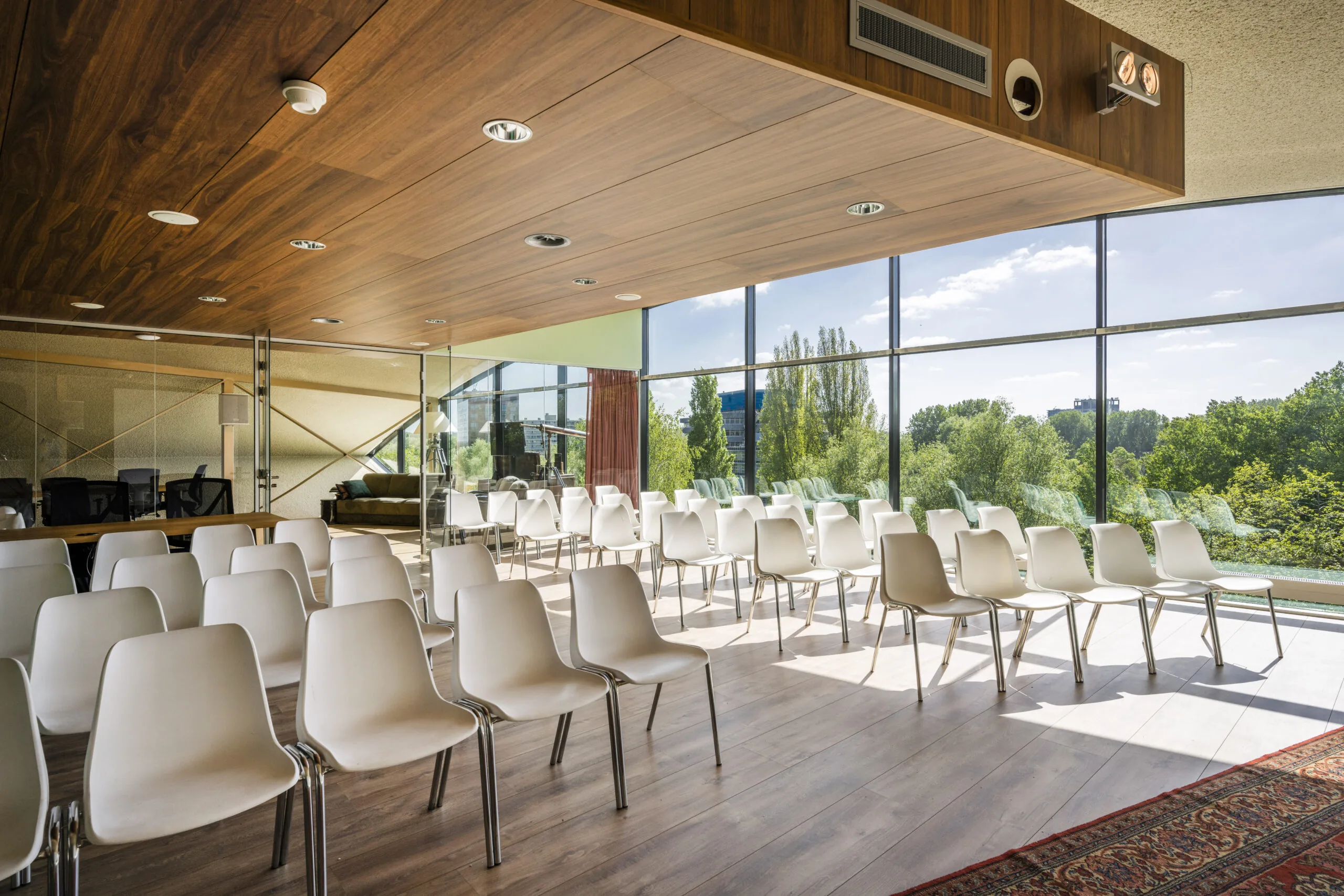 Zaal met witte stoelen en een houten plafond, omgeven door grote ramen met uitzicht op groen.