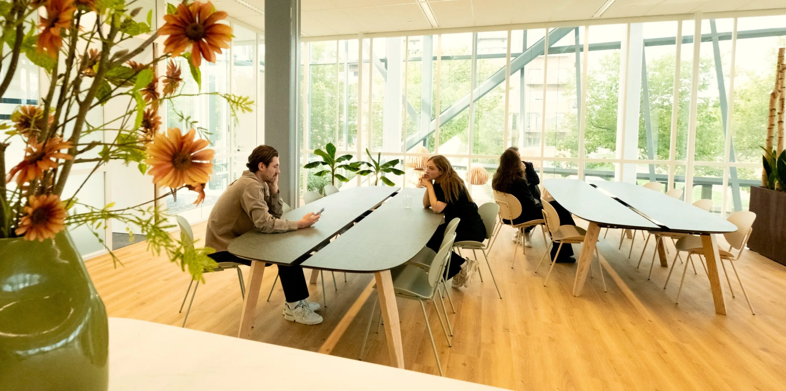 Twee mensen zitten aan een tafel in een moderne kantoor-keuken met veel daglicht en groen.