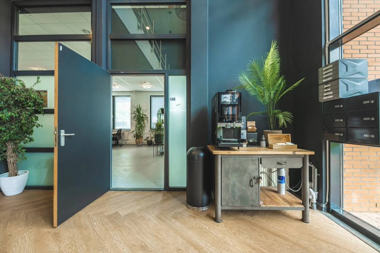 Groot koffiezetapparaat op een metalen tafel in een moderne kantooromgeving met planten en postvakken.