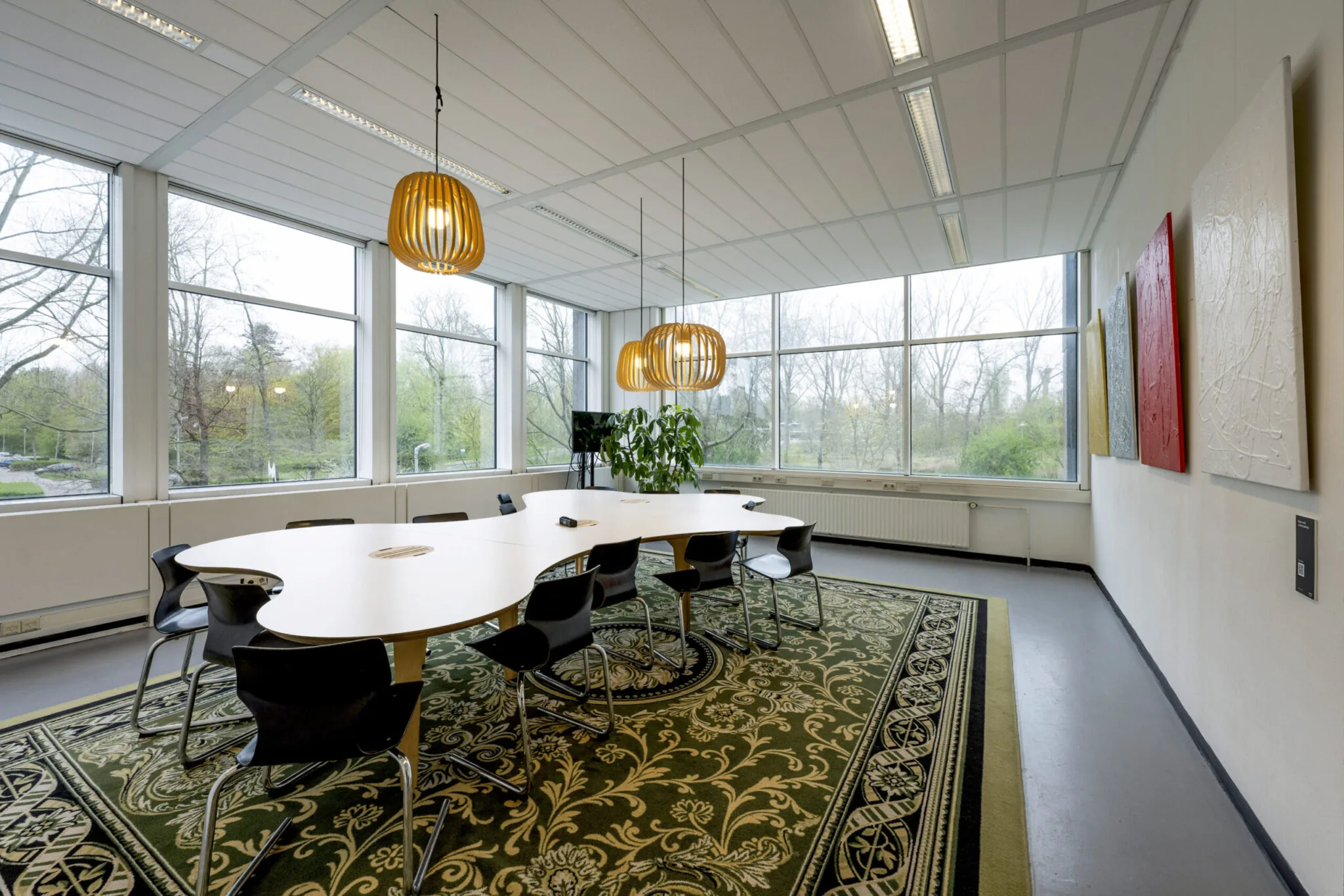 Vergaderruimte Kronenburg 018 met een golvend conferentiebureau, moderne hanglampen, kunst van Paul Starink aan de muur en uitzicht op een groene buitenomgeving.