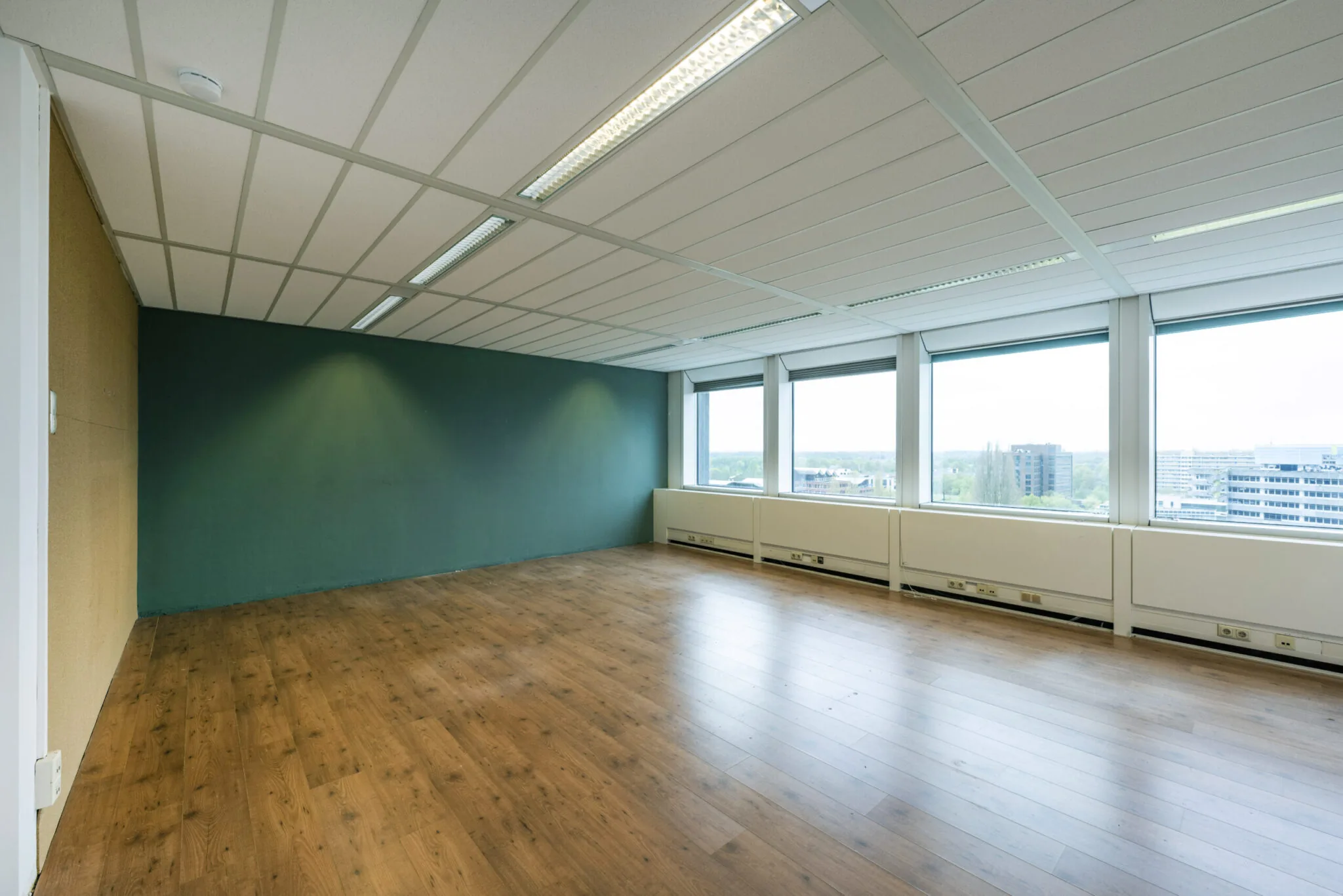 Licht en leeg kantoorpand in Kronenburg met houten vloer, groene accentmuur en uitzicht op de stad.