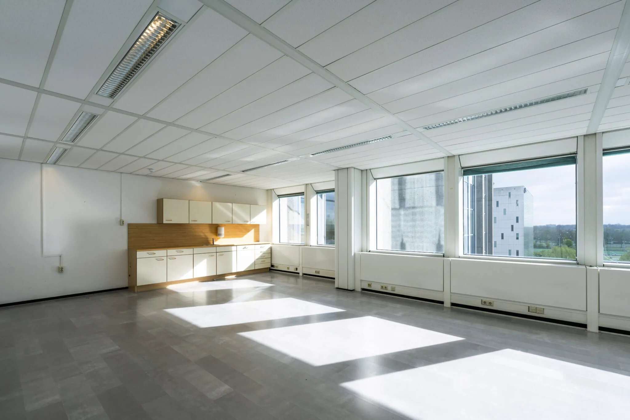 Lichte, lege kantoorruimte in Kronenburg 040 met een eenvoudige kitchenette en grote ramen met uitzicht op een modern kantoorgebouw.