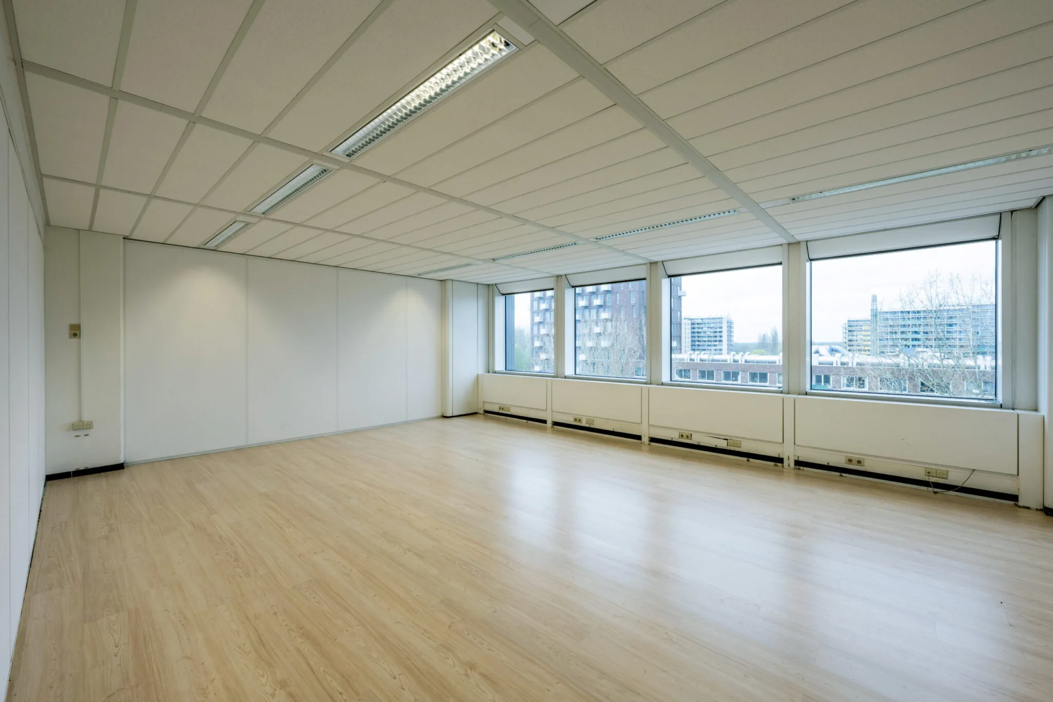 Lege kantoorruimte met licht houten vloer, systeemplafond en grote ramen met uitzicht op flats in Kronenburg.