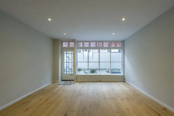 Lichte, lege woonkamer in de Leliestraat met houten vloer en grote ramen met glas-in-lood bovenlichten.