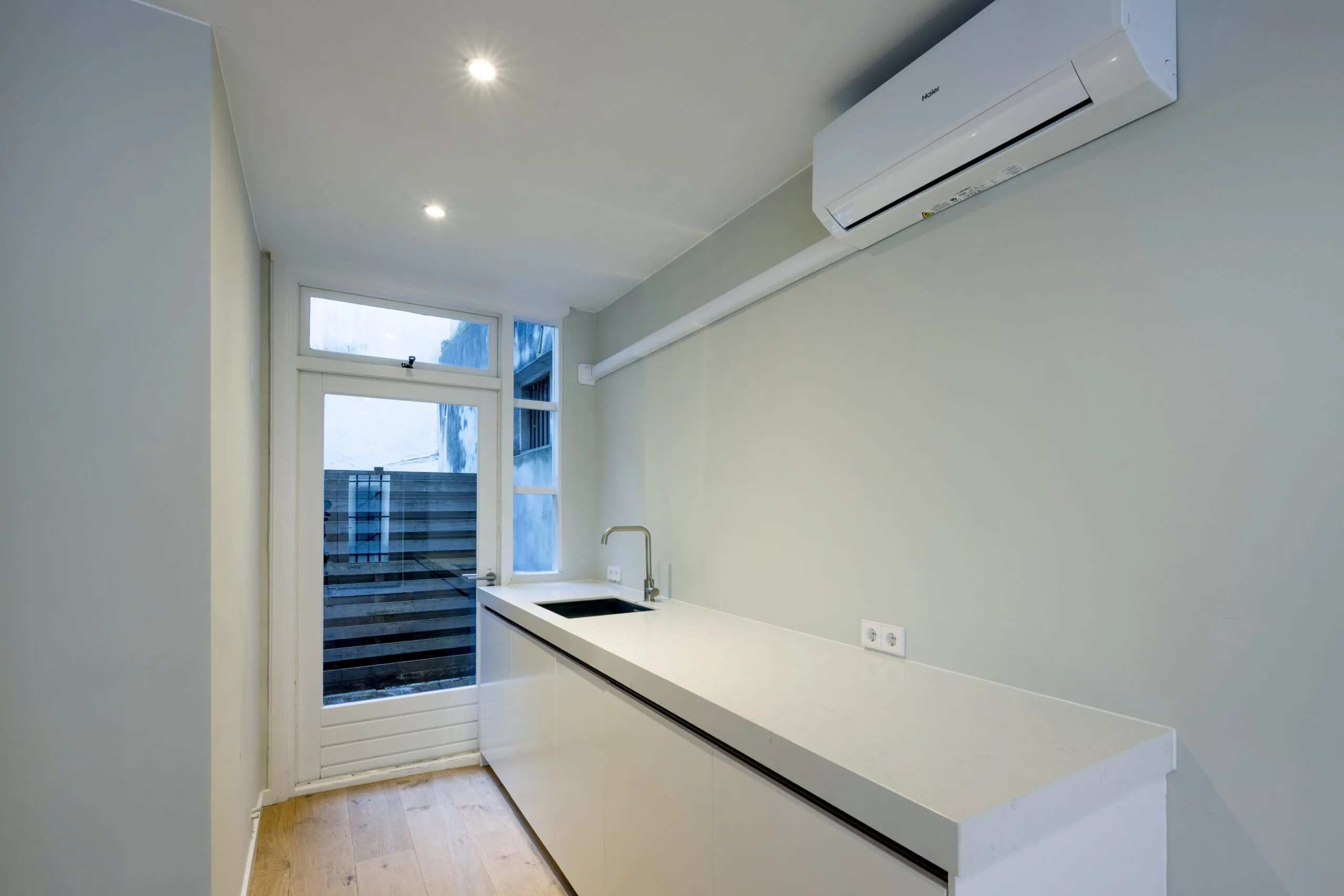 Strakke, moderne keuken in Leliestraat 016 met wit aanrechtblad, ingebouwde spoelbak en airconditioning aan de muur.