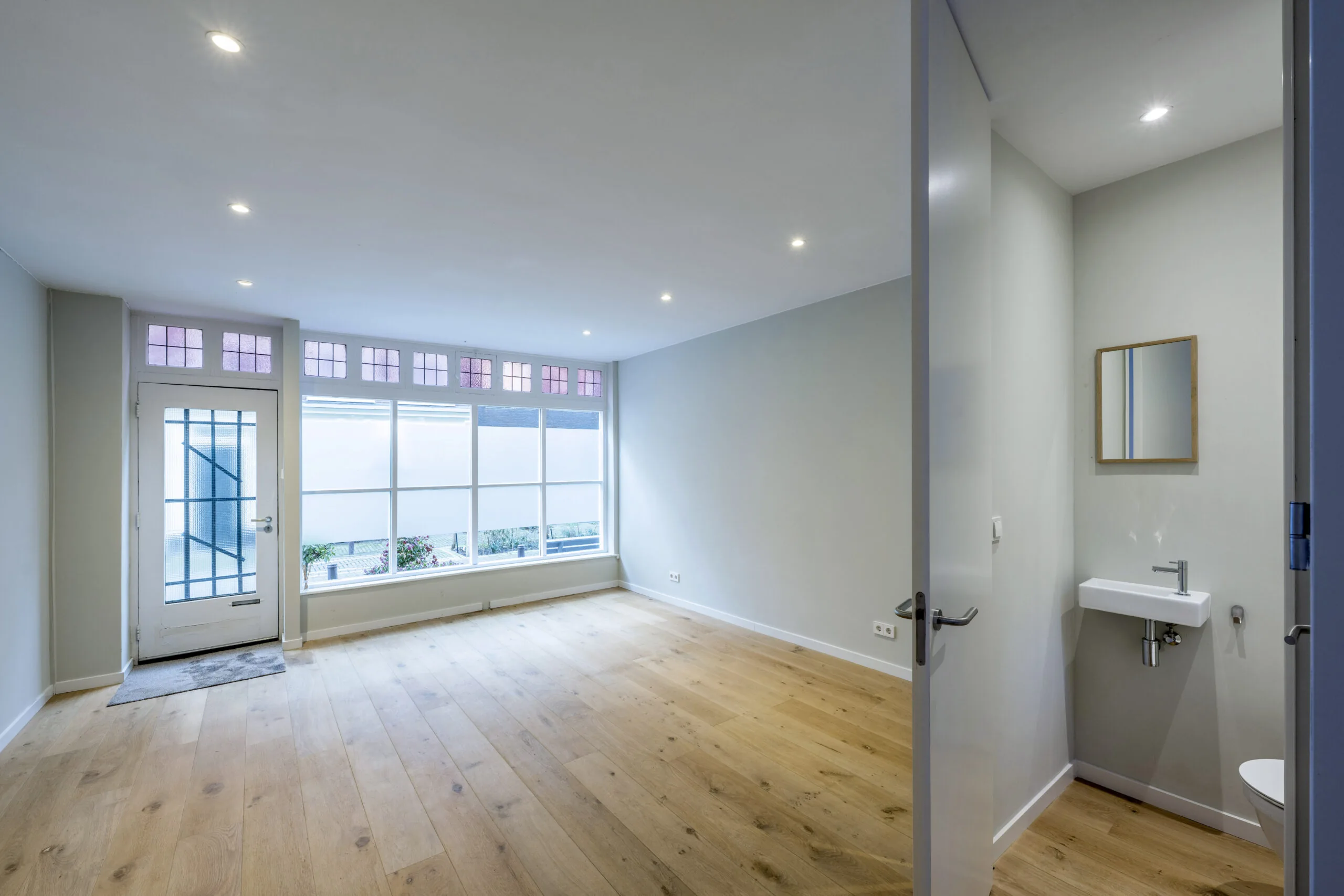 Lichte woonkamer aan de Leliestraat met houten vloer, grote ramen en aangrenzend toilet met wastafel.