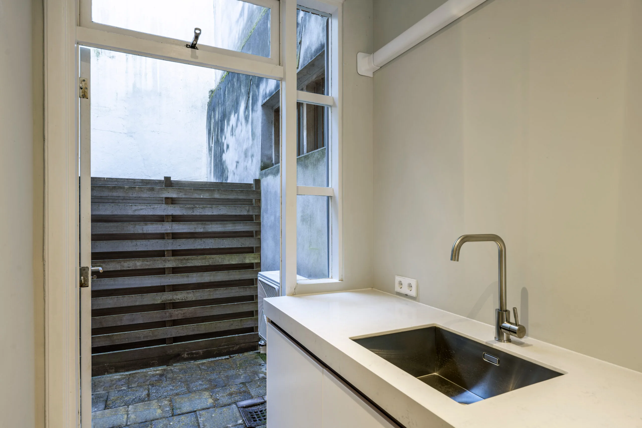 Keuken met wit aanrecht en rvs-kraan, grenzend aan een kleine betegelde buitenruimte met houten schutting, in woning aan Leliestraat 023 van Paul Starink.