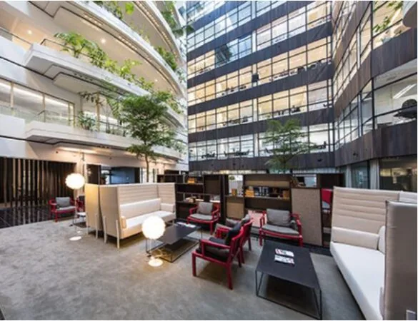 Een moderne lounge met witte banken, rode stoelen en veel planten in een ruim, glazen gebouw.