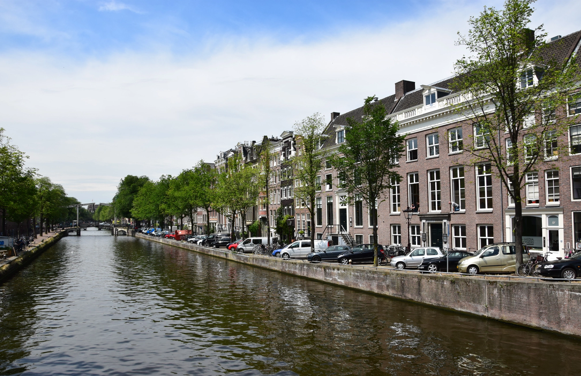 Uitzicht op de Nieuwe Herengracht in Amsterdam met grachtenpanden, geparkeerde auto's en bomen langs het water.
