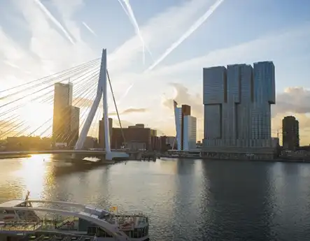 Skyline of Rotterdam at sunset featuring the Erasmus Bridge and modern high-rise buildings along the river.