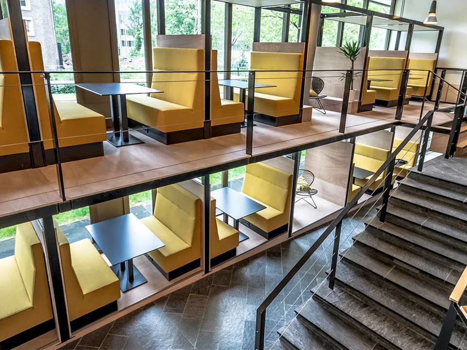 Moderne kantoorruimte met gele zitbanken, tafels en glazen scheidingswanden op twee verdiepingen.