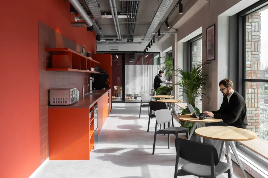 Een moderne, open kantoorruimte met een felrode keuken en een man die aan een tafel bij de ramen werkt.