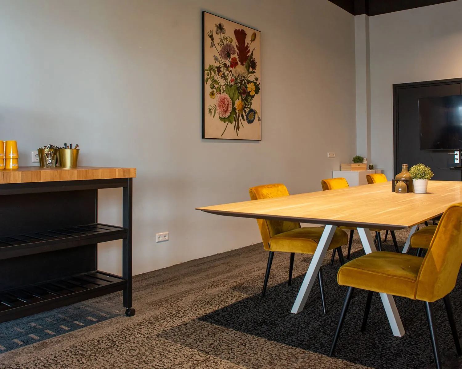 Vergaderruimte met een houten tafel, gele stoelen, een bloemenkunstwerk aan de muur en een bijzettafel met koffiebenodigdheden.