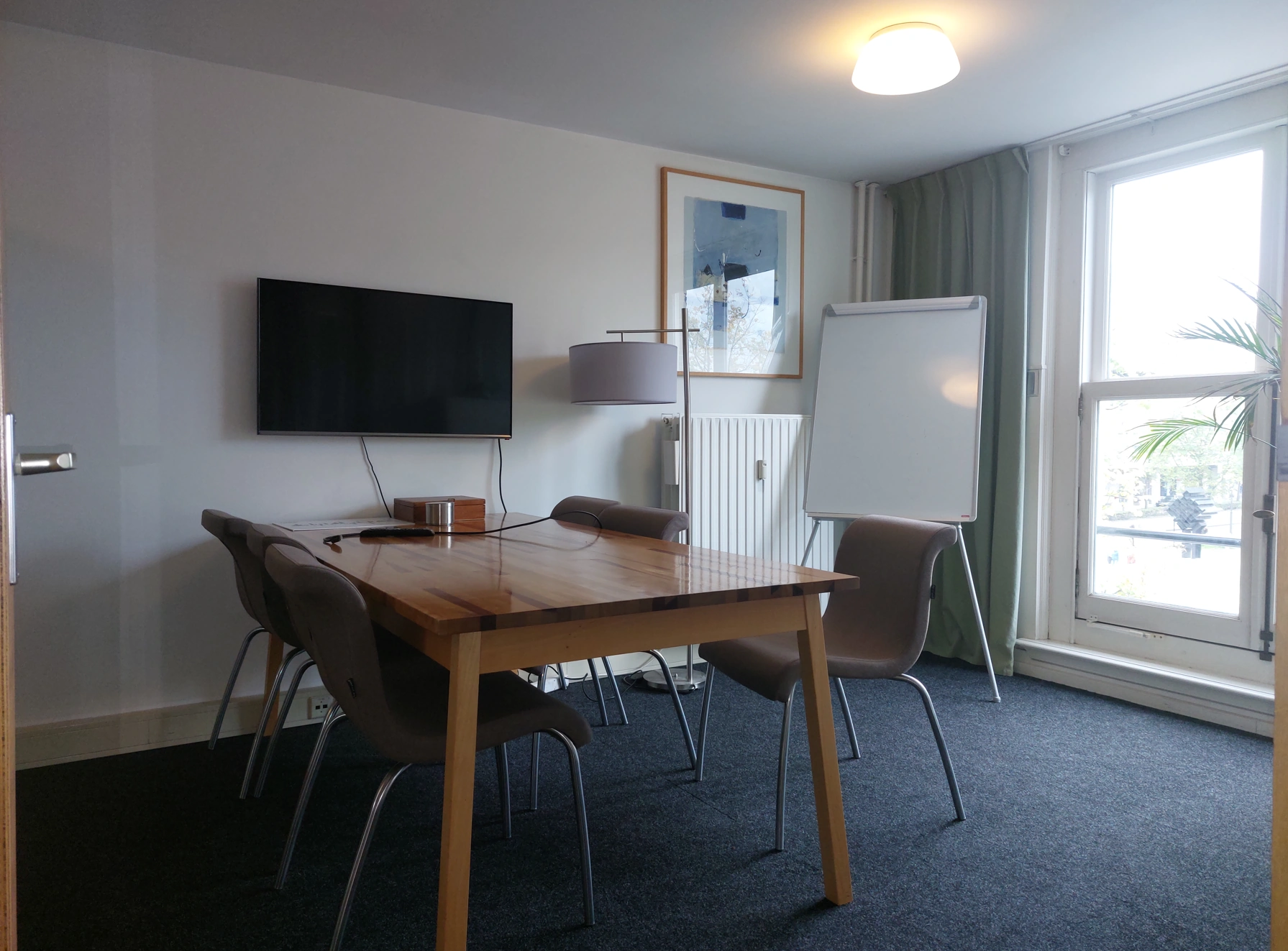 Vergaderruimte met houten tafel, zes stoelen, een flatscreen, een whiteboard en een groot raam.