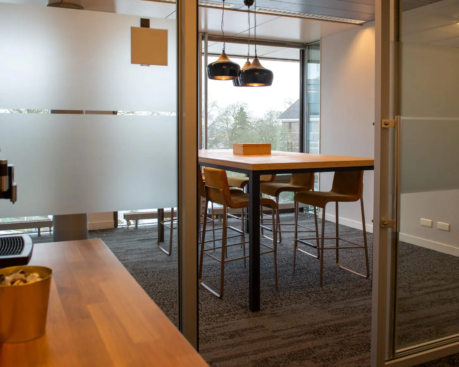 Vergaderruimte met hoge tafel, vier barkrukken en moderne hanglampen bij grote ramen.